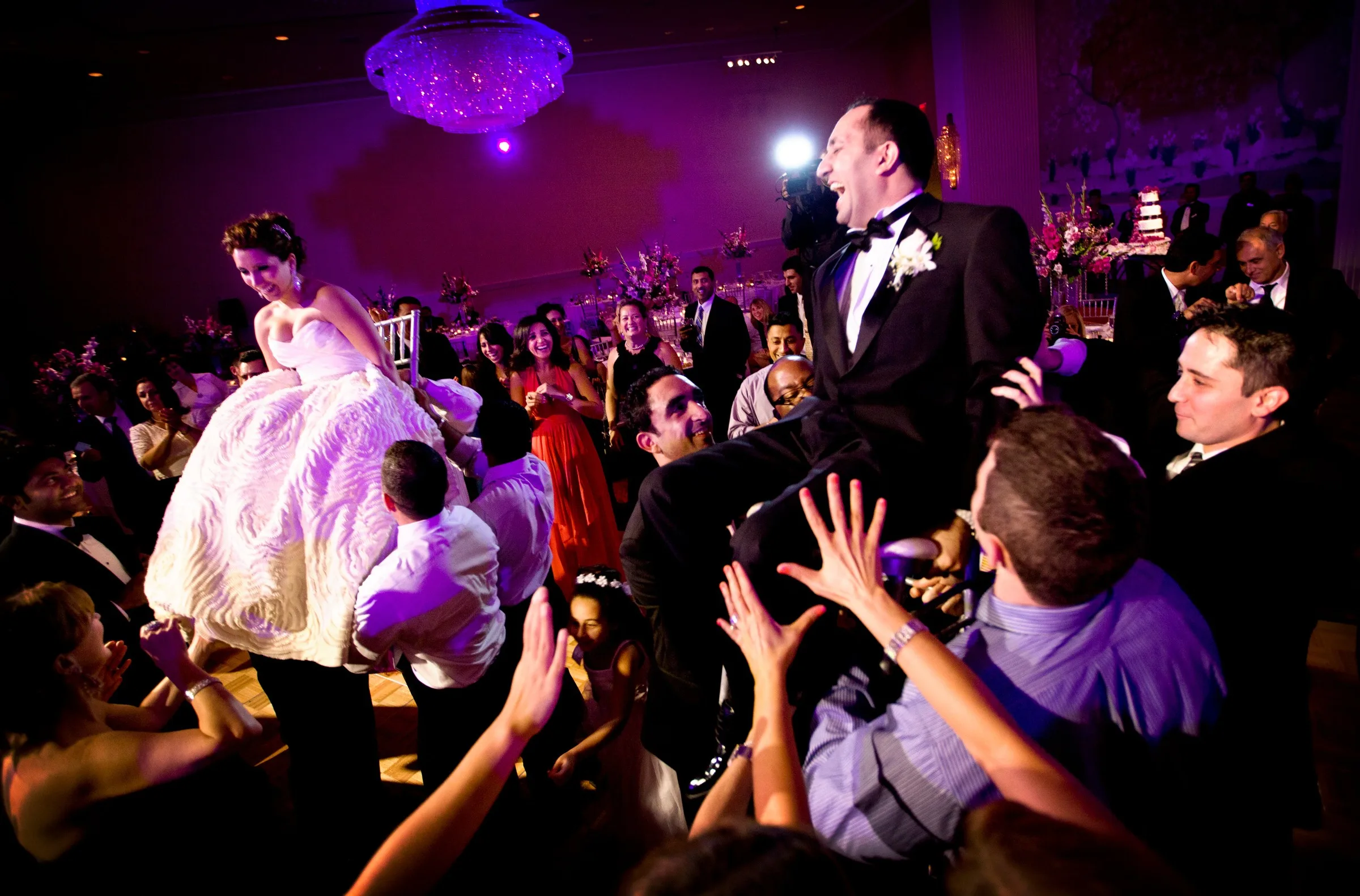 Bride and groom lifted in chairs during the hora at a lively wedding reception in New England, with guests cheering around them on the dance floor