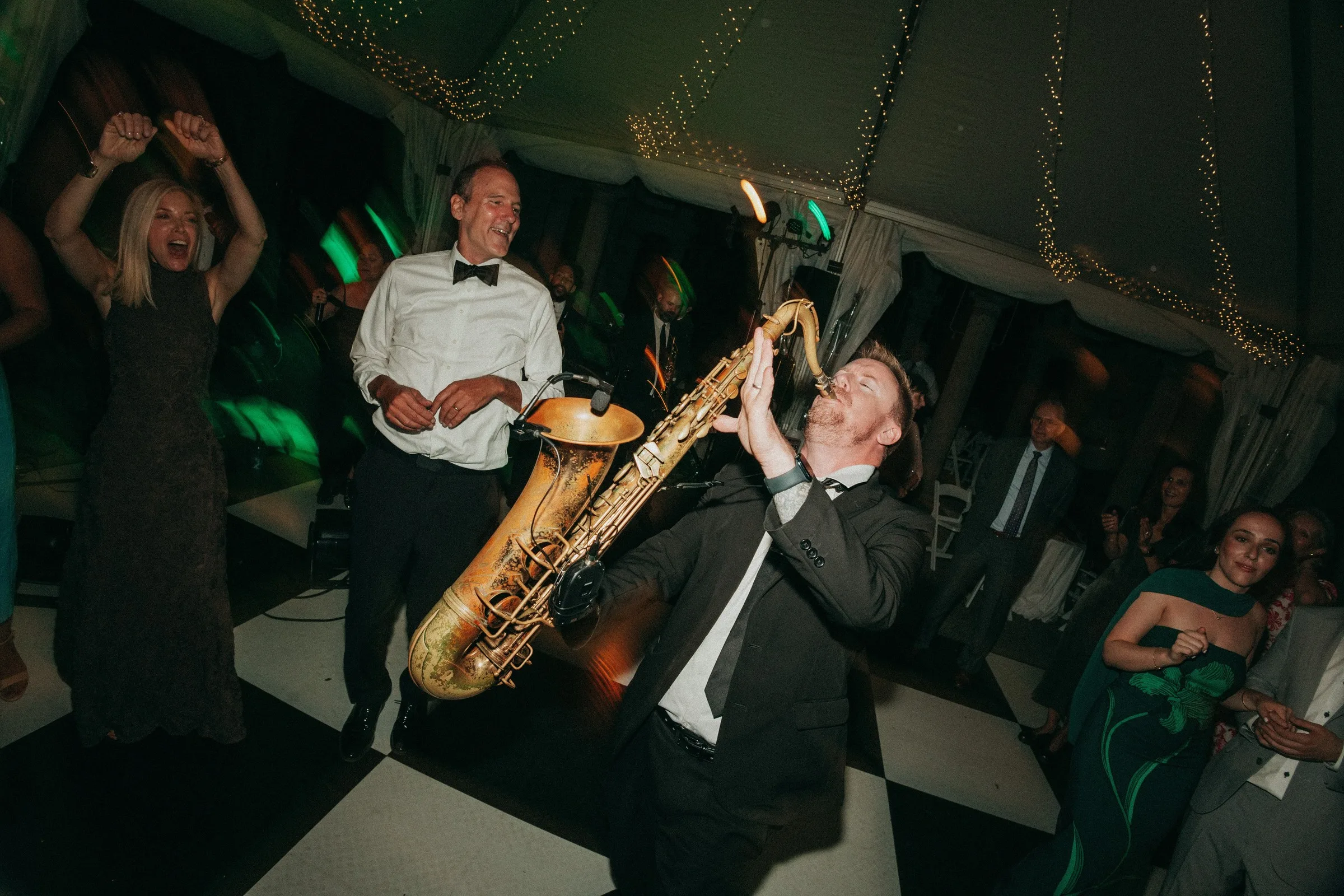 Soul City Band saxophonist performs energetically on a black and white checkered dance floor as guests celebrate at a tented wedding reception in Newport RI