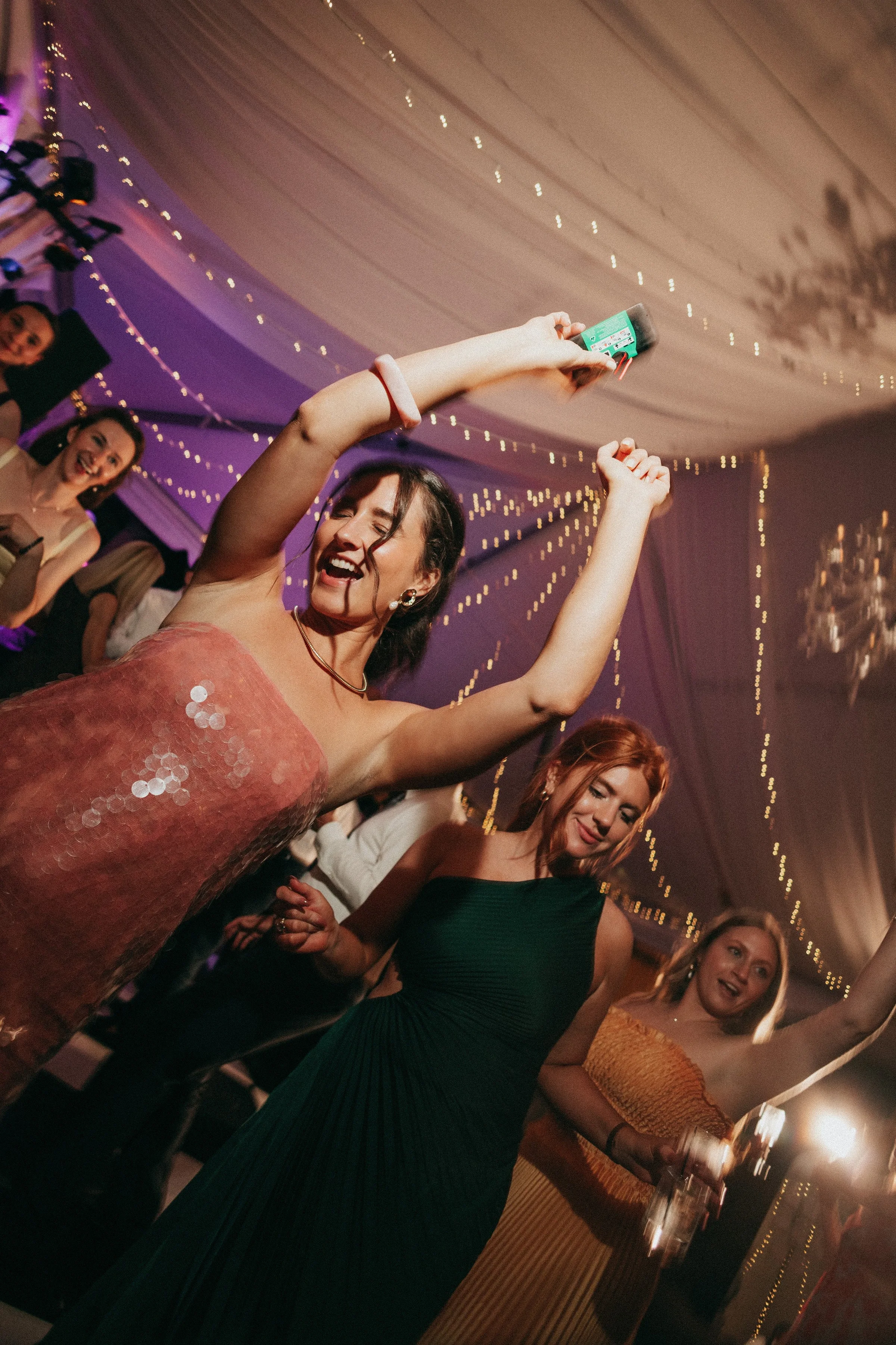 Guests dancing and celebrating with arms raised at a tented wedding reception with fairy lights, energized by live band entertainment in Newport RI