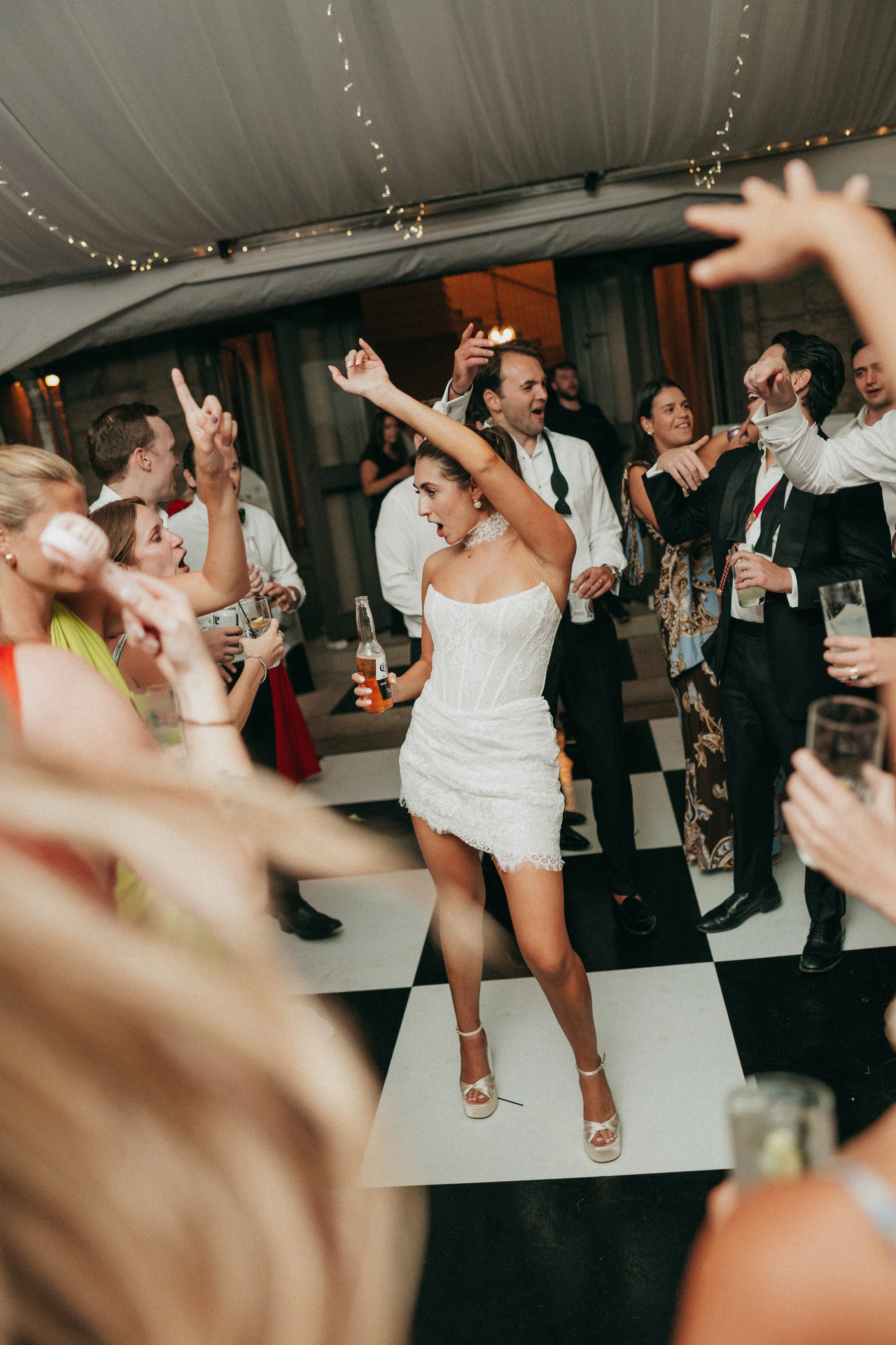 Bride in a short white lace dress dancing energetically on a black and white checkered dance floor surrounded by cheering wedding guests at a tented reception, with live entertainment by Soul City Band in Newport RI