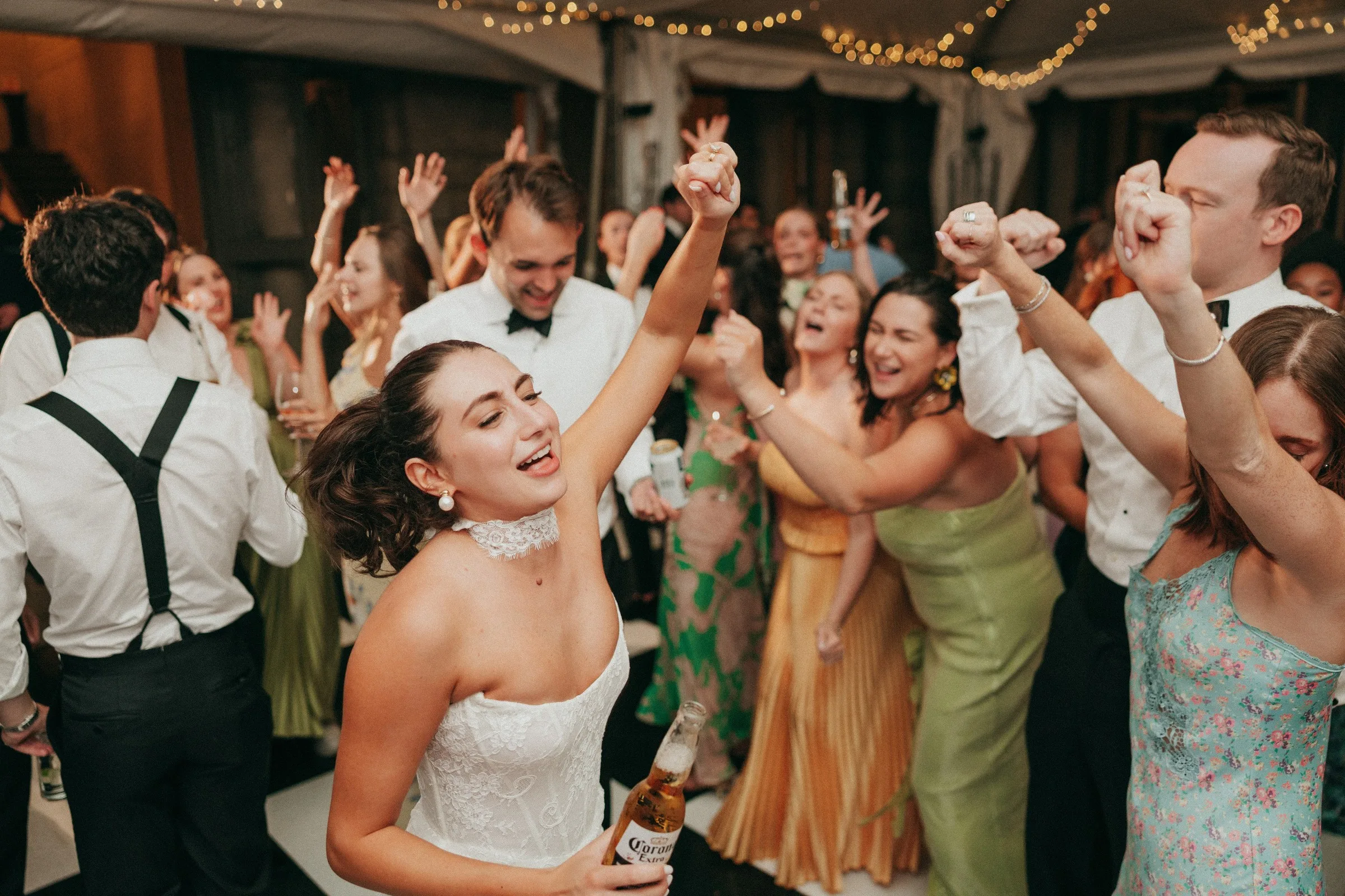 Bride in white strapless gown dancing with raised arm and Corona beer surrounded by joyful wedding guests at a lively reception, captured at a Newport Rhode Island wedding featuring Soul City Band live music entertainment